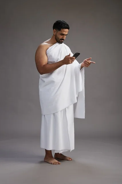 A portrait of an Arab Saudi Gulf man wearing Ihram clothing using a mobile phone, concept of Hajj and Umrah, full body, gray background.