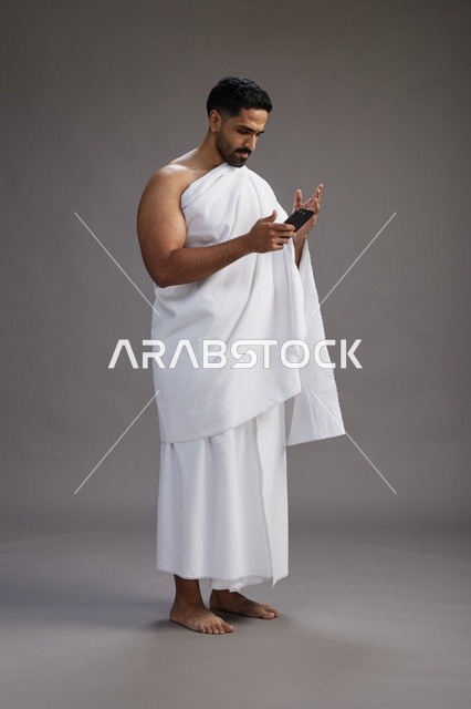 بورتريه لرجل عربي سعودي خليجي يرتدي ملابس الإحرام يستخدم الهاتف المحمول, مفهوم الحج والعمره , كامل الجسم, خلفيه رماديه