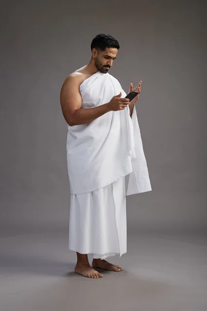 A portrait of an Arab Saudi Gulf man wearing Ihram clothing using a mobile phone, concept of Hajj and Umrah, full body, gray background.
