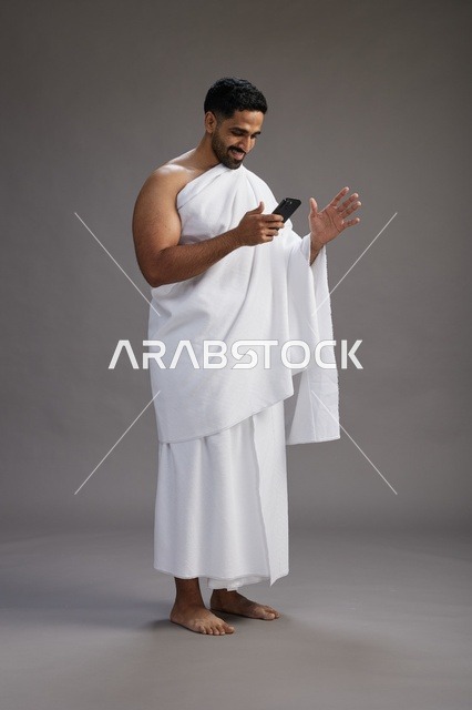 بورتريه لرجل عربي سعودي خليجي يرتدي ملابس الإحرام يستخدم الهاتف المحمول, مفهوم الحج والعمره , كامل الجسم, خلفيه رماديه