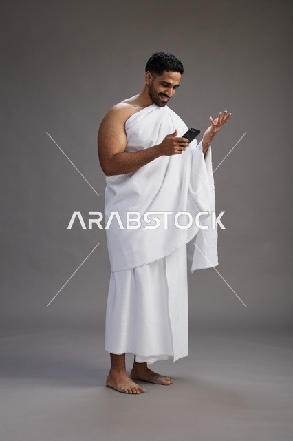 بورتريه لرجل عربي سعودي خليجي يرتدي ملابس الإحرام يستخدم الهاتف المحمول, مفهوم الحج والعمره , كامل الجسم, خلفيه رماديه