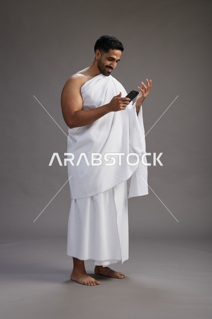 بورتريه لرجل عربي سعودي خليجي يرتدي ملابس الإحرام يستخدم الهاتف المحمول, مفهوم الحج والعمره , كامل الجسم, خلفيه رماديه
