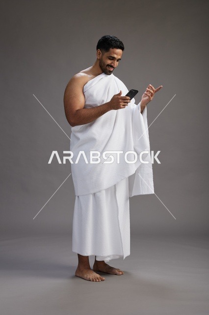 بورتريه لرجل عربي سعودي خليجي يرتدي ملابس الإحرام يستخدم الهاتف المحمول, مفهوم الحج والعمره , كامل الجسم, خلفيه رماديه