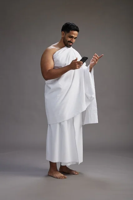 A portrait of an Arab Saudi Gulf man wearing Ihram clothing using a mobile phone, concept of Hajj and Umrah, full body, gray background.