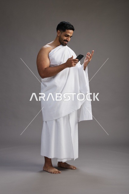 بورتريه لرجل عربي سعودي خليجي يرتدي ملابس الإحرام يستخدم الهاتف المحمول, مفهوم الحج والعمره , كامل الجسم, خلفيه رماديه