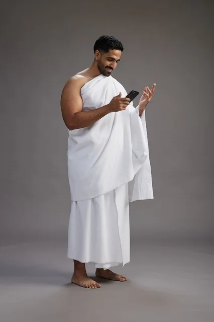 A portrait of an Arab Saudi Gulf man wearing Ihram clothing using a mobile phone, concept of Hajj and Umrah, full body, gray background.
