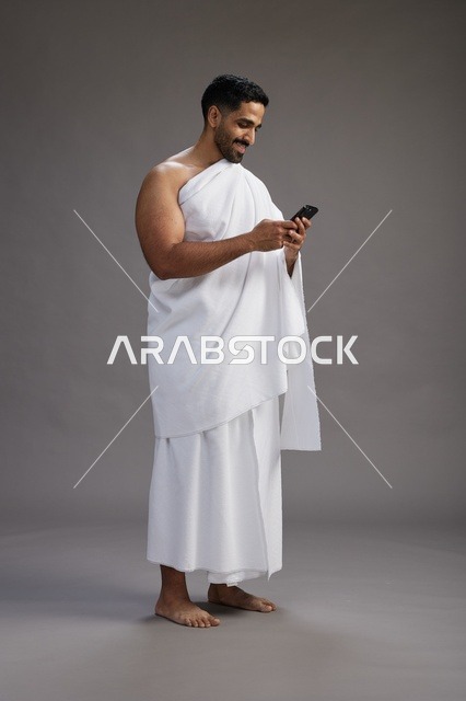 بورتريه لرجل عربي سعودي خليجي يرتدي ملابس الإحرام يستخدم الهاتف المحمول, مفهوم الحج والعمره , كامل الجسم, خلفيه رماديه
