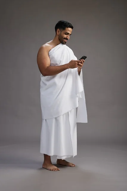 A portrait of an Arab Saudi Gulf man wearing Ihram clothing using a mobile phone, concept of Hajj and Umrah, full body, gray background.