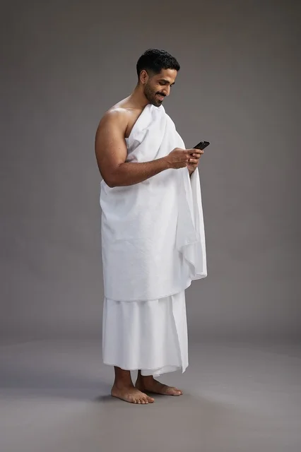 بورتريه لرجل عربي سعودي خليجي يرتدي ملابس الإحرام يستخدم الهاتف المحمول, مفهوم الحج والعمره , كامل الجسم, خلفيه رماديه