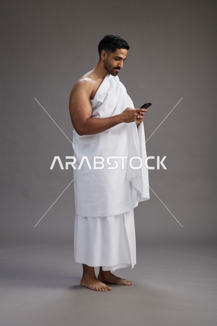 A portrait of an Arab Saudi Gulf man wearing Ihram clothing using a mobile phone, concept of Hajj and Umrah, full body, gray background.
