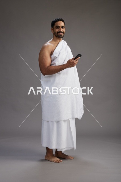 بورتريه لرجل عربي سعودي خليجي يرتدي ملابس الإحرام يستخدم الهاتف المحمول, مفهوم الحج والعمره , كامل الجسم, خلفيه رماديه