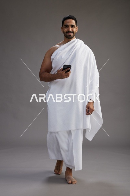 بورتريه لرجل عربي سعودي خليجي يرتدي ملابس الإحرام يستخدم الهاتف المحمول, مفهوم الحج والعمره , كامل الجسم, خلفيه رماديه