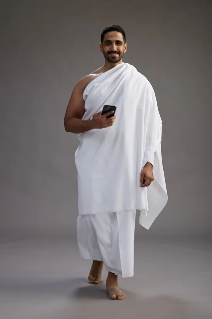 بورتريه لرجل عربي سعودي خليجي يرتدي ملابس الإحرام يستخدم الهاتف المحمول, مفهوم الحج والعمره , كامل الجسم, خلفيه رماديه