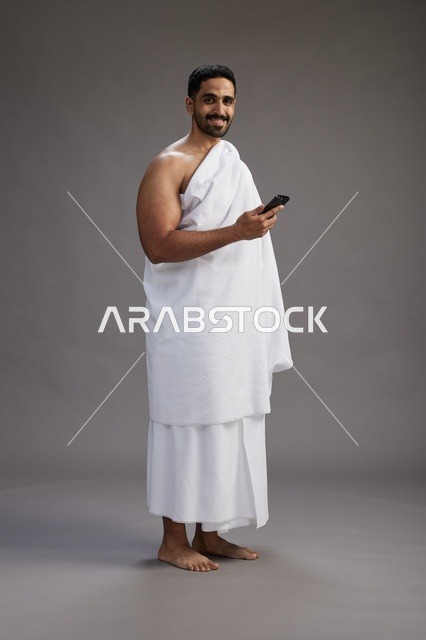 بورتريه لرجل عربي سعودي خليجي يرتدي ملابس الإحرام يستخدم الهاتف المحمول, مفهوم الحج والعمره , كامل الجسم, خلفيه رماديه
