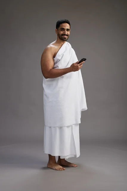 A portrait of an Arab Saudi Gulf man wearing Ihram clothing using a mobile phone, concept of Hajj and Umrah, full body, gray background.