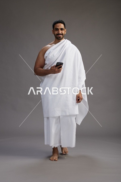 بورتريه لرجل عربي سعودي خليجي يرتدي ملابس الإحرام يستخدم الهاتف المحمول, مفهوم الحج والعمره , كامل الجسم, خلفيه رماديه