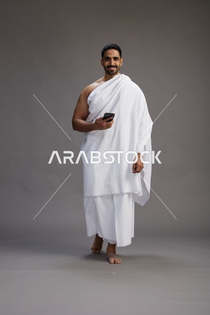 بورتريه لرجل عربي سعودي خليجي يرتدي ملابس الإحرام يستخدم الهاتف المحمول, مفهوم الحج والعمره , كامل الجسم, خلفيه رماديه