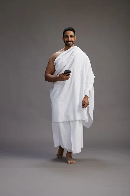 A portrait of an Arab Saudi Gulf man wearing Ihram clothing using a mobile phone, concept of Hajj and Umrah, full body, gray background.