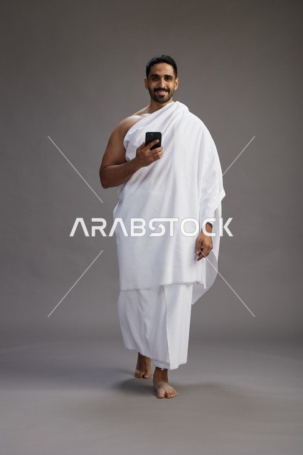 A portrait of an Arab Saudi Gulf man wearing Ihram clothing using a mobile phone, concept of Hajj and Umrah, full body, gray background.