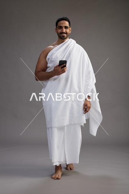 بورتريه لرجل عربي سعودي خليجي يرتدي ملابس الإحرام يستخدم الهاتف المحمول, مفهوم الحج والعمره , كامل الجسم, خلفيه رماديه