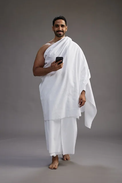 A portrait of an Arab Saudi Gulf man wearing Ihram clothing using a mobile phone, concept of Hajj and Umrah, full body, gray background.