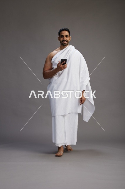 بورتريه لرجل عربي سعودي خليجي يرتدي ملابس الإحرام يستخدم الهاتف المحمول, مفهوم الحج والعمره , كامل الجسم, خلفيه رماديه