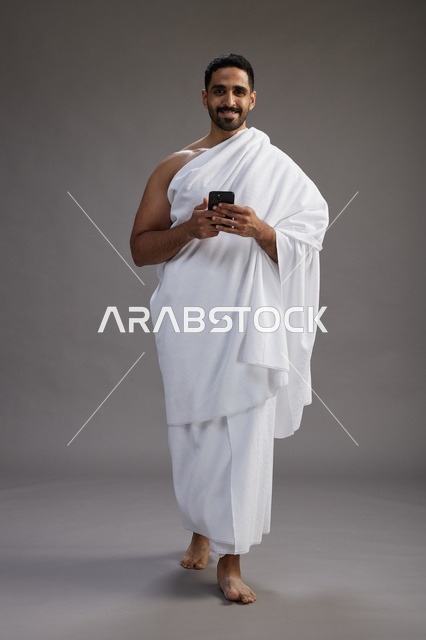 بورتريه لرجل عربي سعودي خليجي يرتدي ملابس الإحرام يستخدم الهاتف المحمول, مفهوم الحج والعمره , كامل الجسم, خلفيه رماديه