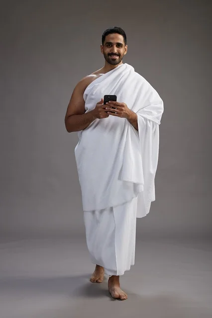 بورتريه لرجل عربي سعودي خليجي يرتدي ملابس الإحرام يستخدم الهاتف المحمول, مفهوم الحج والعمره , كامل الجسم, خلفيه رماديه