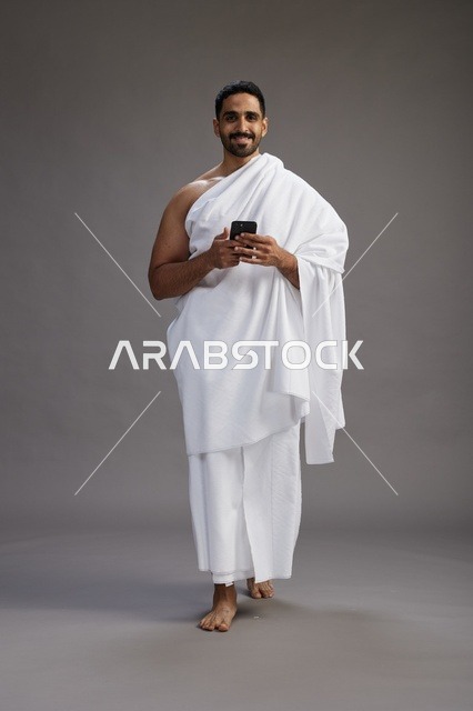بورتريه لرجل عربي سعودي خليجي يرتدي ملابس الإحرام يستخدم الهاتف المحمول, مفهوم الحج والعمره , كامل الجسم, خلفيه رماديه