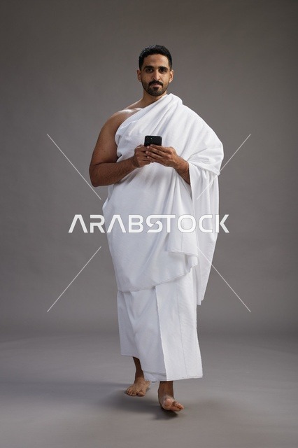 بورتريه لرجل عربي سعودي خليجي يرتدي ملابس الإحرام يستخدم الهاتف المحمول, مفهوم الحج والعمره , كامل الجسم, خلفيه رماديه