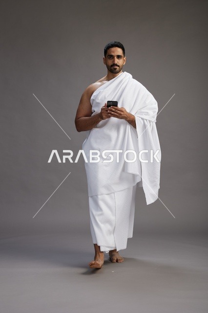 بورتريه لرجل عربي سعودي خليجي يرتدي ملابس الإحرام يستخدم الهاتف المحمول, مفهوم الحج والعمره , كامل الجسم, خلفيه رماديه