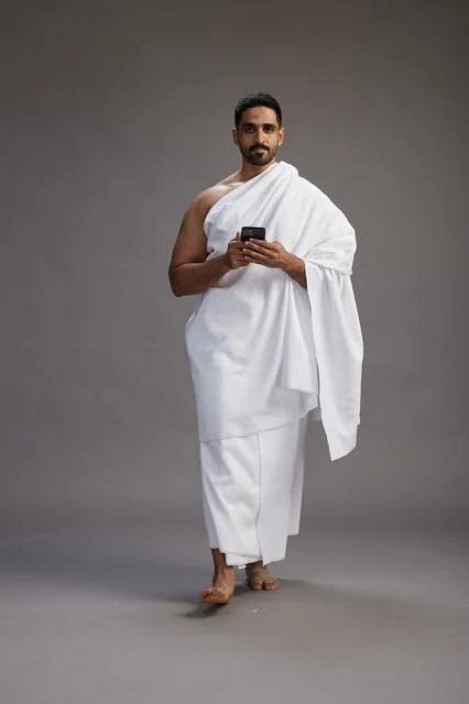 A portrait of an Arab Saudi Gulf man wearing Ihram clothing using a mobile phone, concept of Hajj and Umrah, full body, gray background.