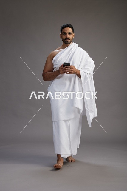 بورتريه لرجل عربي سعودي خليجي يرتدي ملابس الإحرام يستخدم الهاتف المحمول, مفهوم الحج والعمره , كامل الجسم, خلفيه رماديه