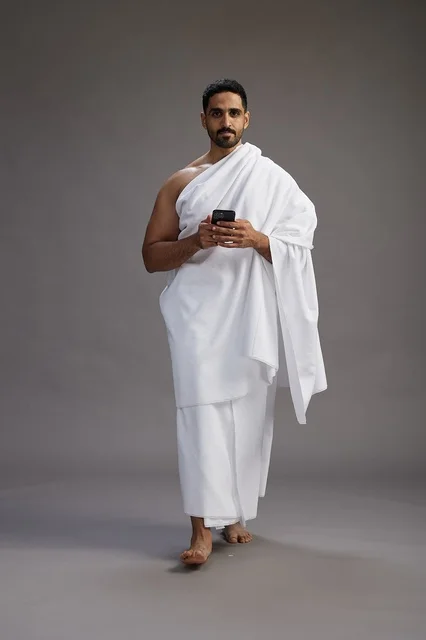A portrait of an Arab Saudi Gulf man wearing Ihram clothing using a mobile phone, concept of Hajj and Umrah, full body, gray background.