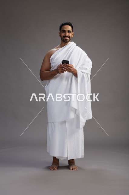 بورتريه لرجل عربي سعودي خليجي يرتدي ملابس الإحرام يستخدم الهاتف المحمول, مفهوم الحج والعمره , كامل الجسم, خلفيه رماديه