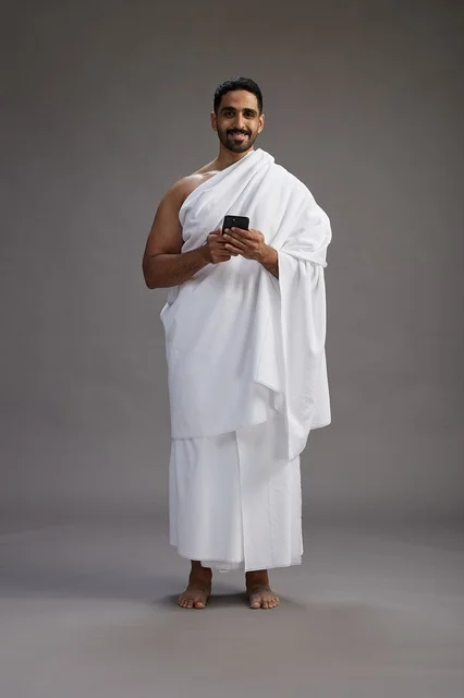 بورتريه لرجل عربي سعودي خليجي يرتدي ملابس الإحرام يستخدم الهاتف المحمول, مفهوم الحج والعمره , كامل الجسم, خلفيه رماديه