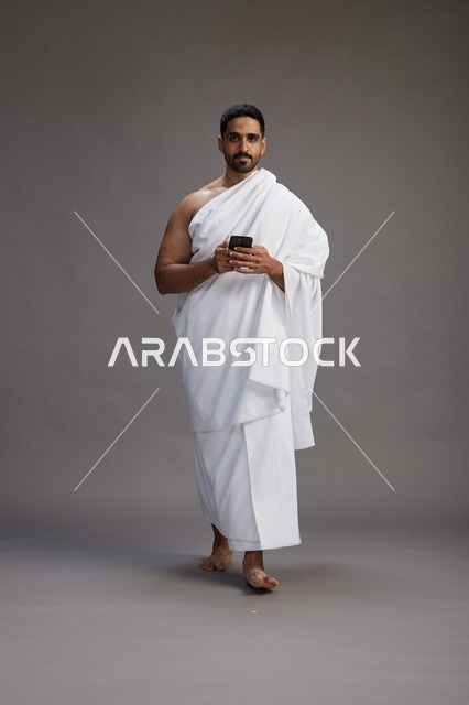 بورتريه لرجل عربي سعودي خليجي يرتدي ملابس الإحرام يستخدم الهاتف المحمول, مفهوم الحج والعمره , كامل الجسم, خلفيه رماديه