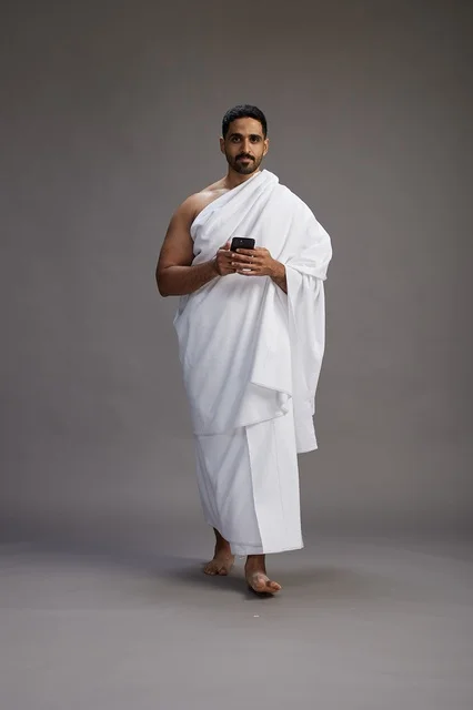 A portrait of an Arab Saudi Gulf man wearing Ihram clothing using a mobile phone, concept of Hajj and Umrah, full body, gray background.