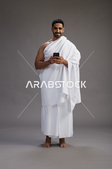 بورتريه لرجل عربي سعودي خليجي يرتدي ملابس الإحرام يستخدم الهاتف المحمول, مفهوم الحج والعمره , كامل الجسم, خلفيه رماديه