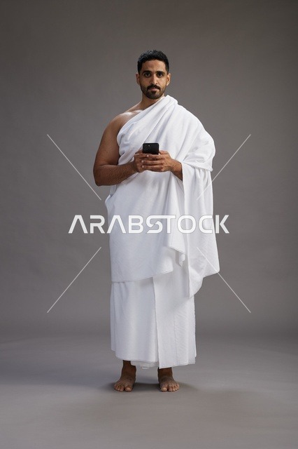 بورتريه لرجل عربي سعودي خليجي يرتدي ملابس الإحرام يستخدم الهاتف المحمول, مفهوم الحج والعمره , كامل الجسم, خلفيه رماديه