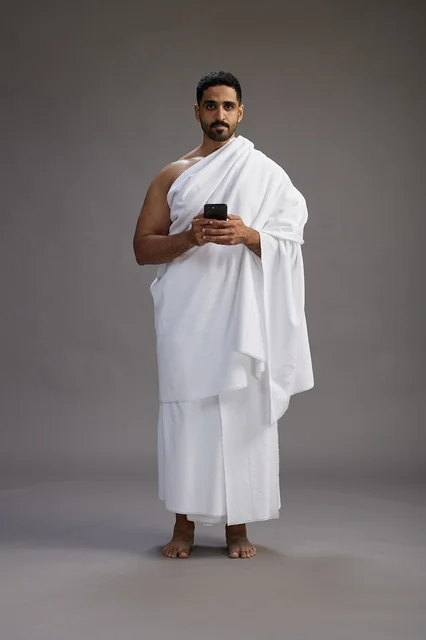 A portrait of an Arab Saudi Gulf man wearing Ihram clothing using a mobile phone, concept of Hajj and Umrah, full body, gray background.