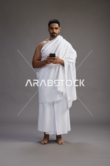 بورتريه لرجل عربي سعودي خليجي يرتدي ملابس الإحرام يستخدم الهاتف المحمول, مفهوم الحج والعمره , كامل الجسم, خلفيه رماديه