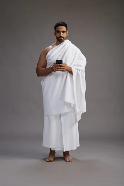 A portrait of an Arab Saudi Gulf man wearing Ihram clothing using a mobile phone, concept of Hajj and Umrah, full body, gray background.