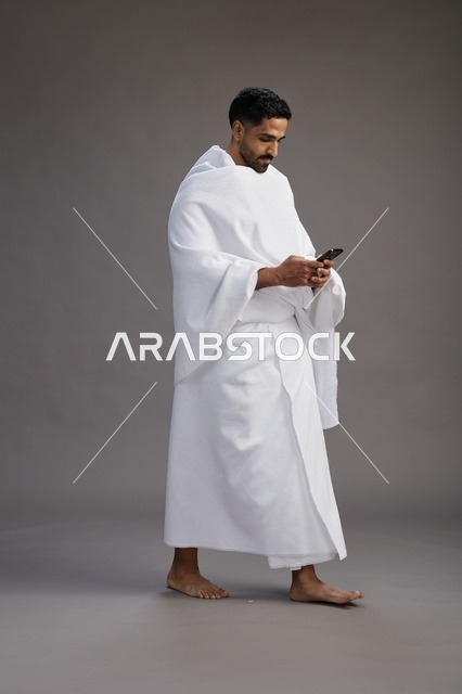 بورتريه لرجل عربي سعودي خليجي يرتدي ملابس الإحرام يستخدم الهاتف المحمول, مفهوم الحج والعمره , كامل الجسم, خلفيه رماديه