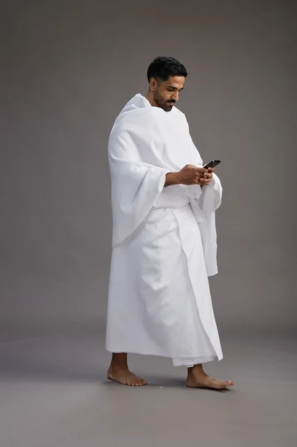 بورتريه لرجل عربي سعودي خليجي يرتدي ملابس الإحرام يستخدم الهاتف المحمول, مفهوم الحج والعمره , كامل الجسم, خلفيه رماديه