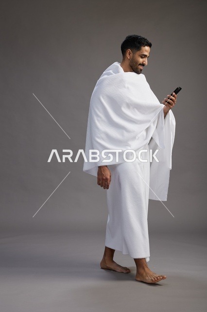 بورتريه لرجل عربي سعودي خليجي يرتدي ملابس الإحرام يستخدم الهاتف المحمول, مفهوم الحج والعمره , كامل الجسم, خلفيه رماديه