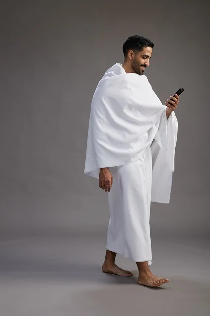 A portrait of an Arab Saudi Gulf man wearing Ihram clothing using a mobile phone, concept of Hajj and Umrah, full body, gray background.