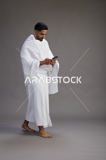 بورتريه لرجل عربي سعودي خليجي يرتدي ملابس الإحرام يستخدم الهاتف المحمول, مفهوم الحج والعمره , كامل الجسم, خلفيه رماديه