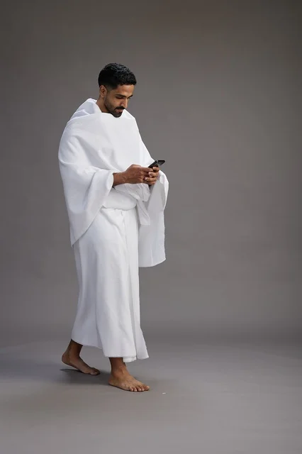 A portrait of an Arab Saudi Gulf man wearing Ihram clothing using a mobile phone, concept of Hajj and Umrah, full body, gray background. A portrait of an Arab Saudi Gulf man wearing Ihram clothing using a mobile phone, concept of Hajj and Umrah, full body, gray background.