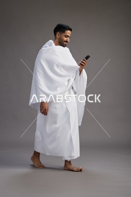بورتريه لرجل عربي سعودي خليجي يرتدي ملابس الإحرام يستخدم الهاتف المحمول, مفهوم الحج والعمره , كامل الجسم, خلفيه رماديه
